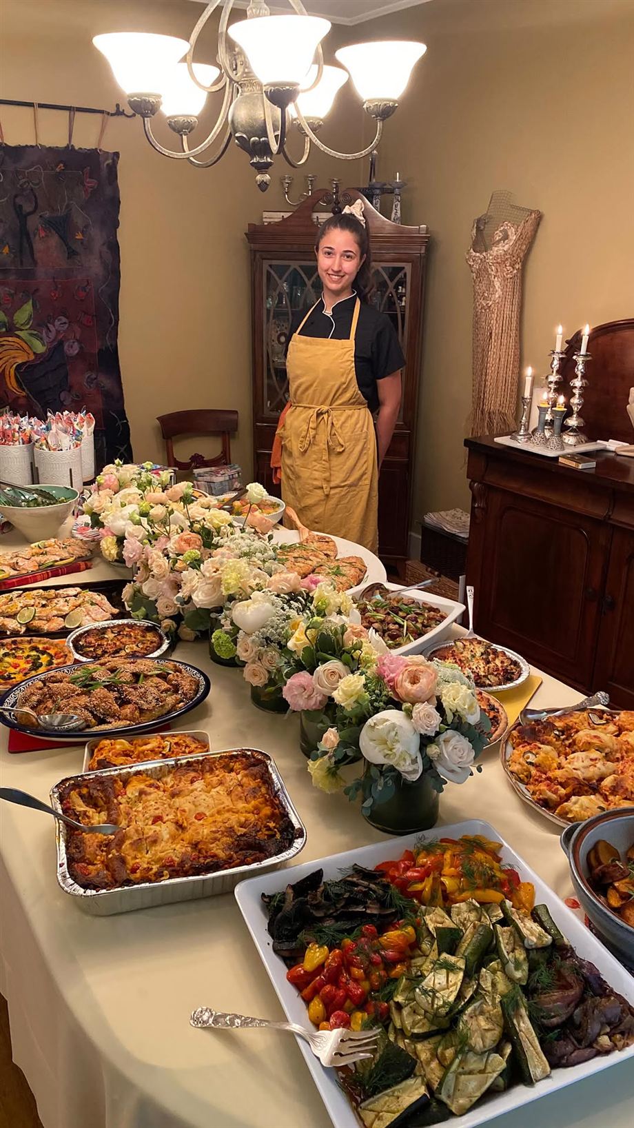 Chef Zoë Rose in her yellow apron standing behind a candlelit table full of platters and flowers at a client's dinner party